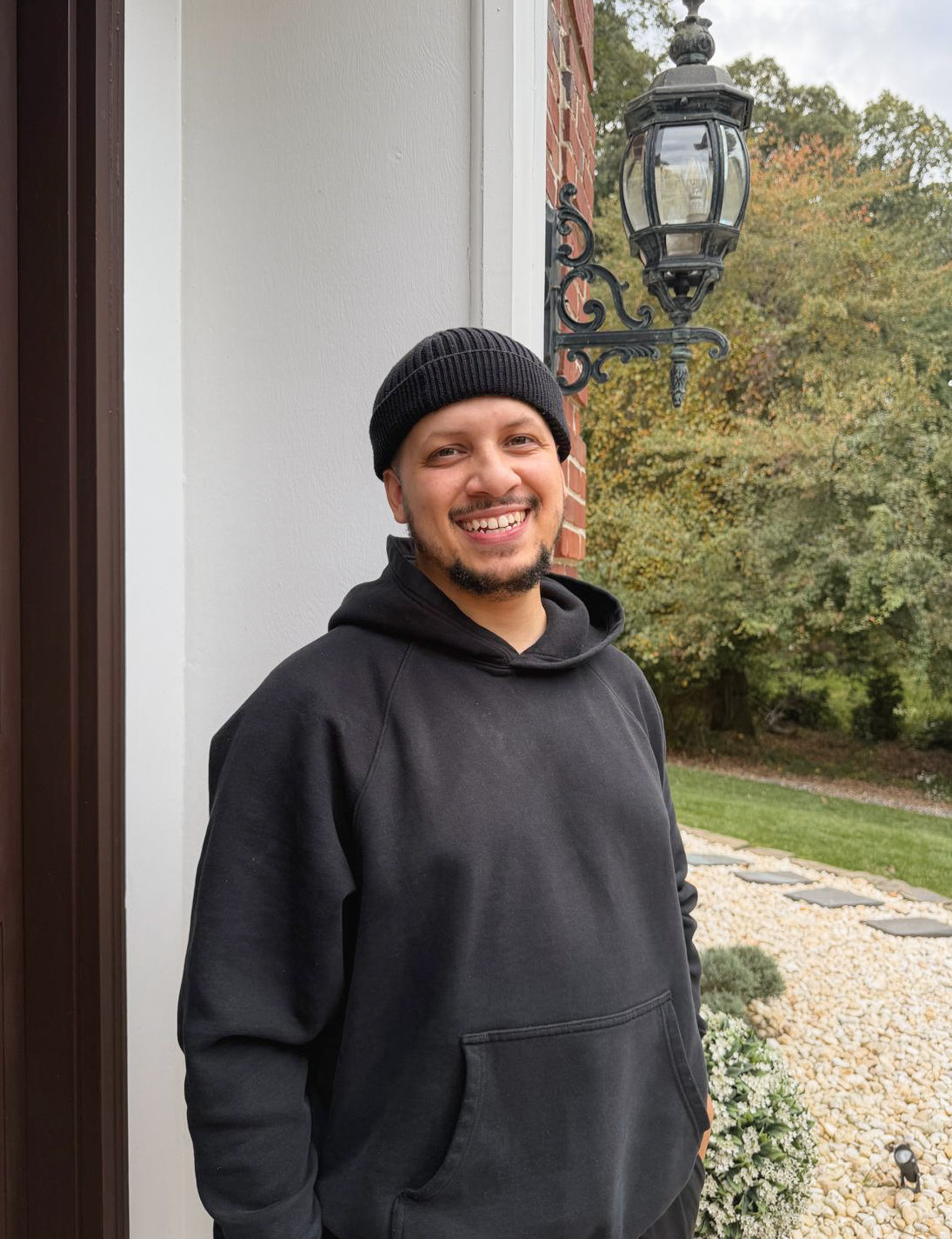 Ayodiran Fabian smiling in a black hoodie and beanie outside a brick home